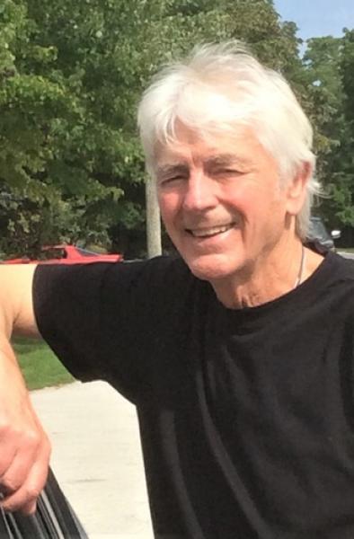 A photo of Allan Davis, smiling and leaning against a car. He has short white hair and is wearing a black T-shirt.