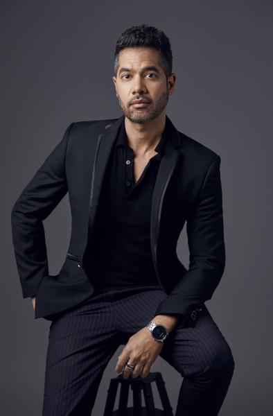 A photo of a man wearing a black suit jacket, black polo shirt, and pinstriped pants. He has brown skin with a bit of facial hair and short hair pushed up and back.