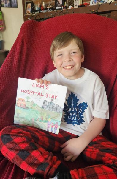 A little boy wearing on a red chair, smiling widely and holding a copy of Liam's Hospital Stay.