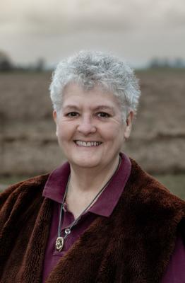 headshot: Ragini Werner. A smiling woman with short grey hair. She is wearing a deep red velvet jacket and a silver necklace, and standing in front of a field.